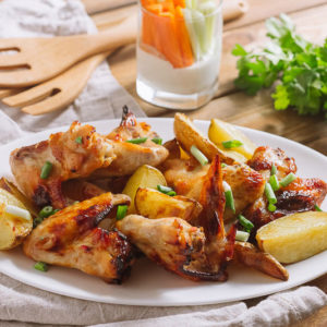 Картофель с овощами и крыльями | Oval white plate with fried chicken wings with fried potatoes. Food sprinkled with chopped scallion. Against the background is a glass with chopped carrots and celery in sour cream. Parsley branch.
