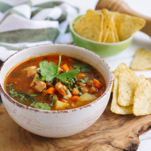 Суп с курицей, нутом и начос | Chicken soup on chicken broth, with nachos, chickpeas and tomatoes in a deep beige bowl. Sprinkled with chopped parsley leaves. On a wooden table, a cutting board, with a pepper mill, a kitchen towel.