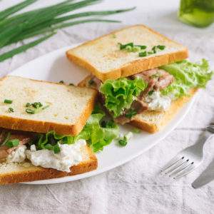 Тосты с свиной шеей и сыром | White wheat bread toasted sandwich with pork meat and cheese, salad and sour cream. Chopped chives, bottled olive oil, a knife and a fork. Against the background of a gray fabric napkin.