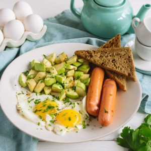 Яйца с сосисками и кабачком | Fried eggs for breakfast on a white plate. Fried eggs with sausages and zucchini, two slices of rye bread, parsley. White wooden background and blue fabric napkin.